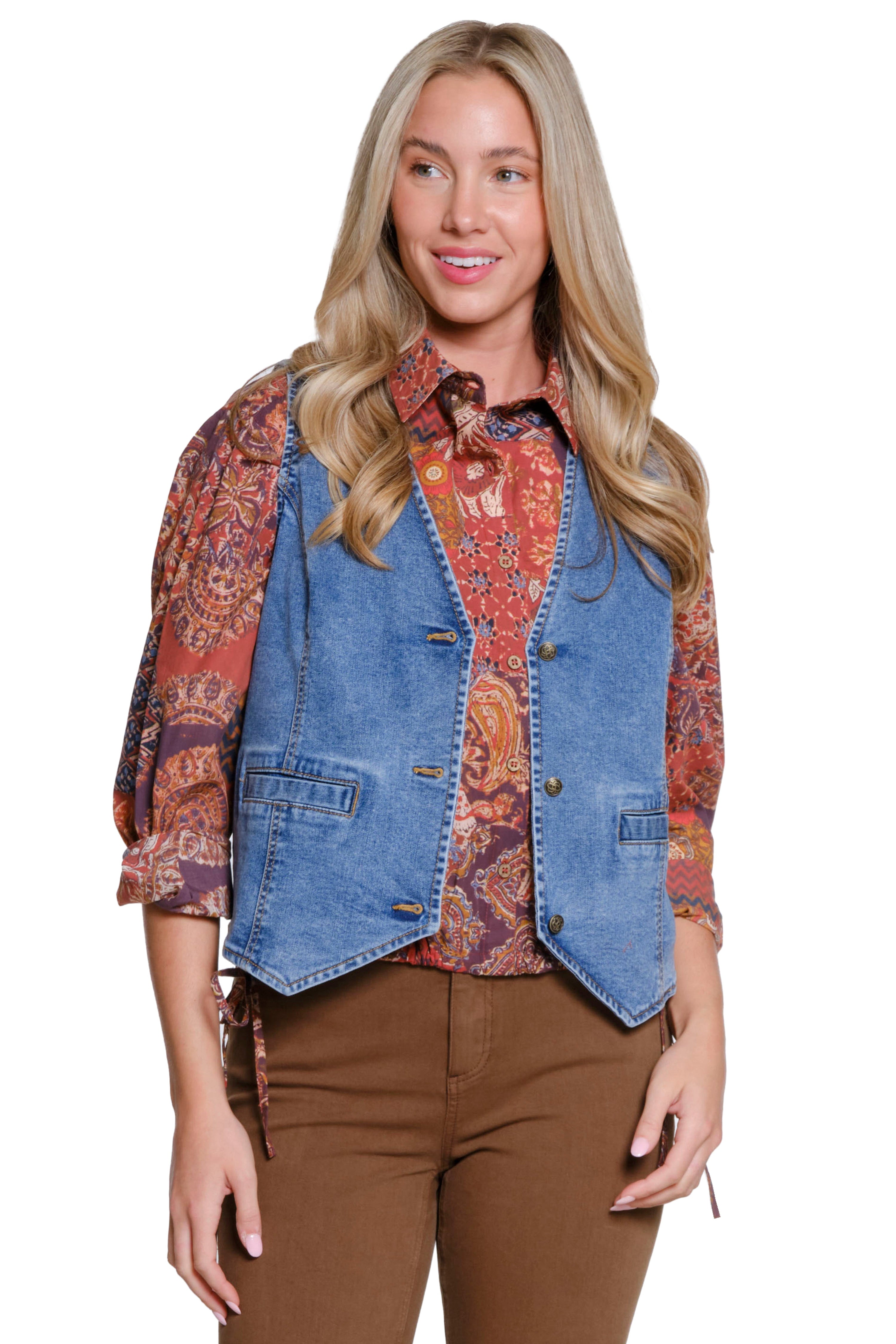Woman wearing a denim vest over a patterned shirt with brown pants on a white background